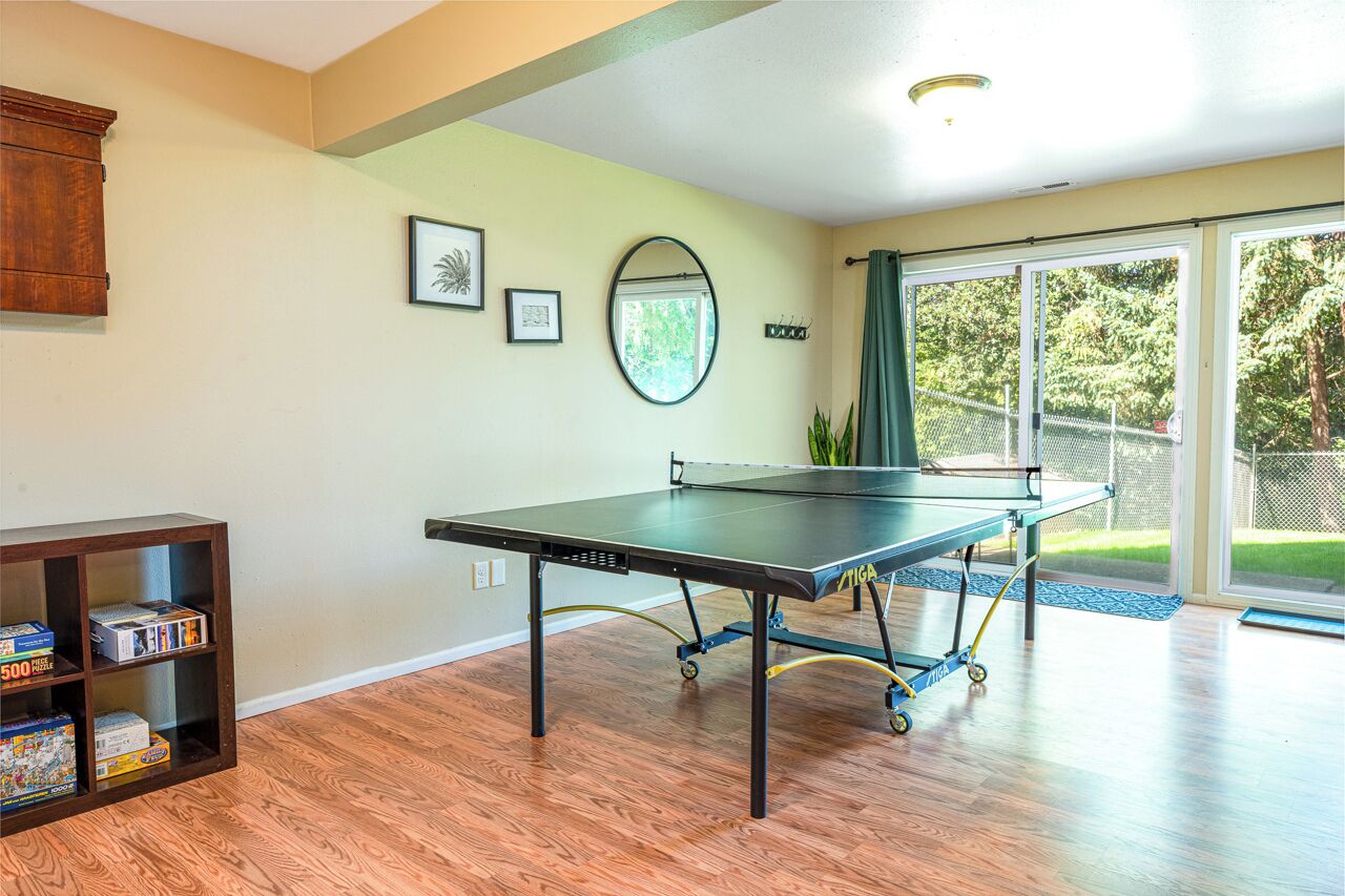 Une table de ping-pong dans une pièce au plancher en bois, un meuble contenant des jeux de société et une grande fenêtre donnant sur un paysage verdoyant.
