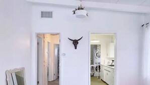 A hallway with a white door, a mirror, a red bench, and a kitchen area visible through an open door. - Pink, Architect-Appointed House! 3 min from JTNP. (TWENTYNINE PALMS)
