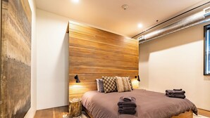 A modern bedroom with a wooden headboard, a large bed with a brown comforter, two bedside lamps, and a window with a view.