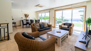 Un'ampia zona giorno con mobili in vimini, un tavolo da pranzo e grandi porte scorrevoli in vetro che si aprono su un balcone con vista sull'oceano.