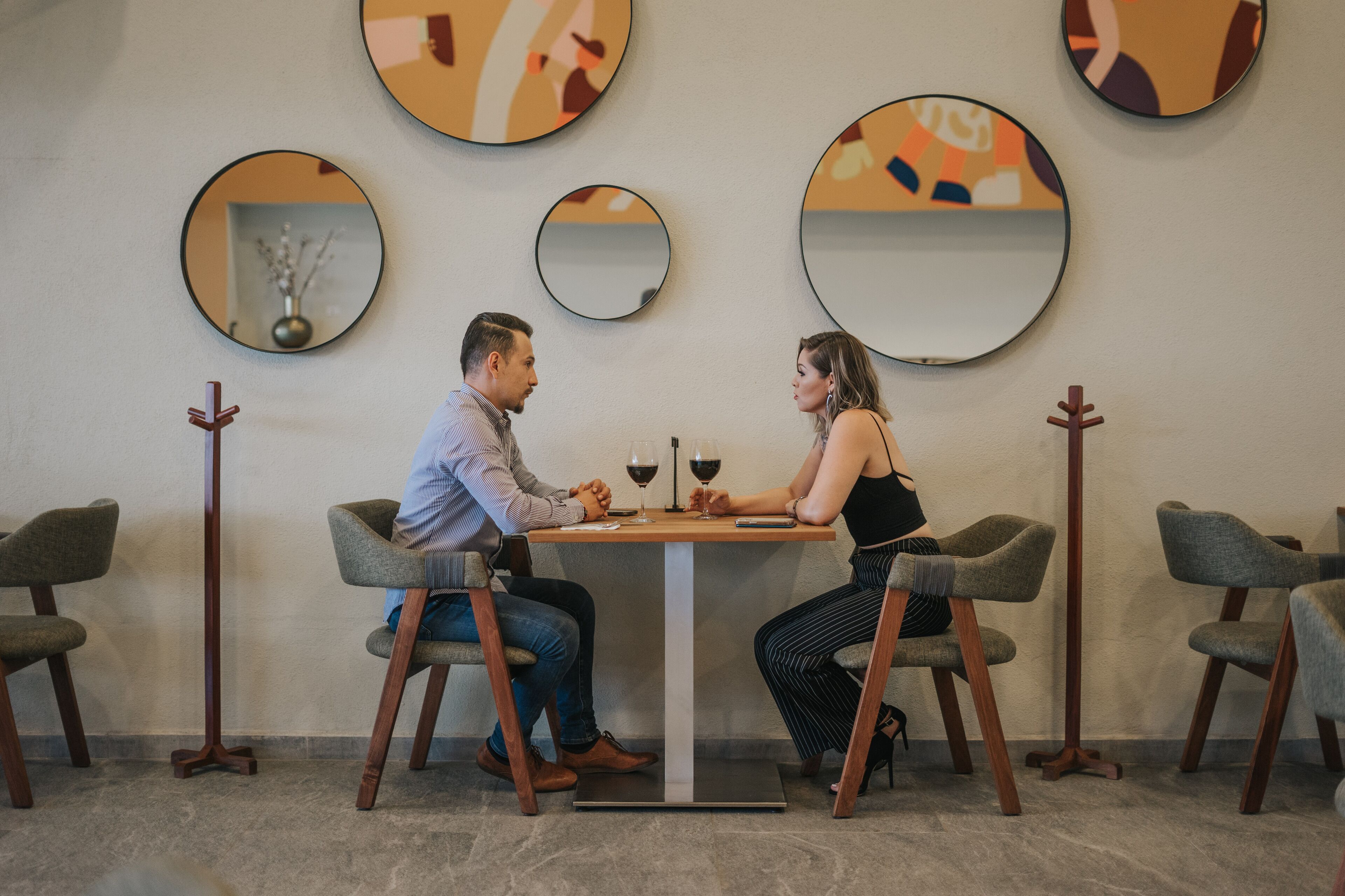Una pareja sentada en una mesa con copas de vino, rodeada de sillas modernas y espejos redondos en la pared.