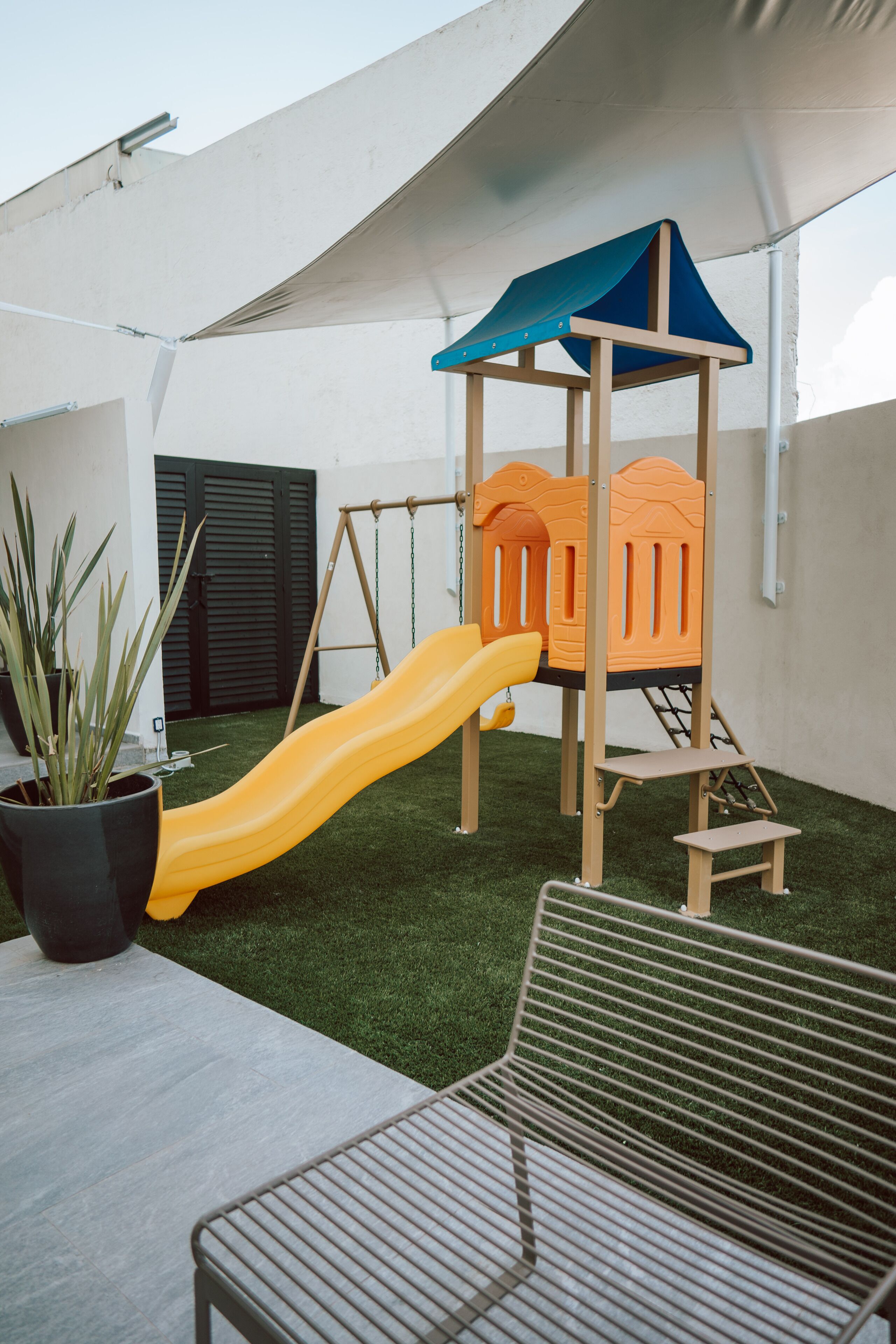 A children's play area with a slide and climbing structure, surrounded by a white wall and a potted plant.