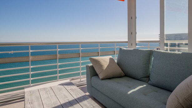 Een balkon met uitzicht op de oceaan, een grijze bank met kussens en een houten terras.