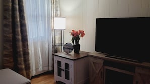 Een kamer met een televisie, een klein tafeltje met een klok en bloemen, een lamp en een raam met gordijnen.
