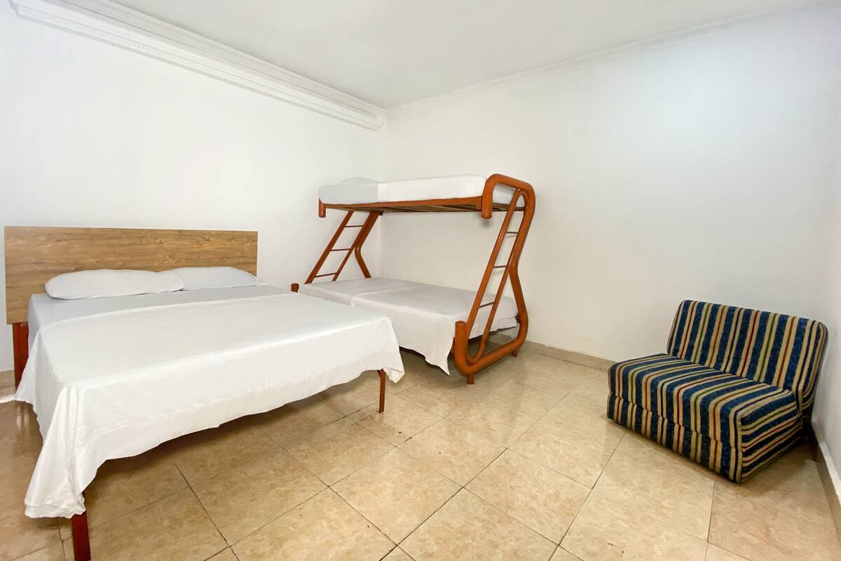 A room with a bunk bed, a single bed, a striped armchair, and a white table.
