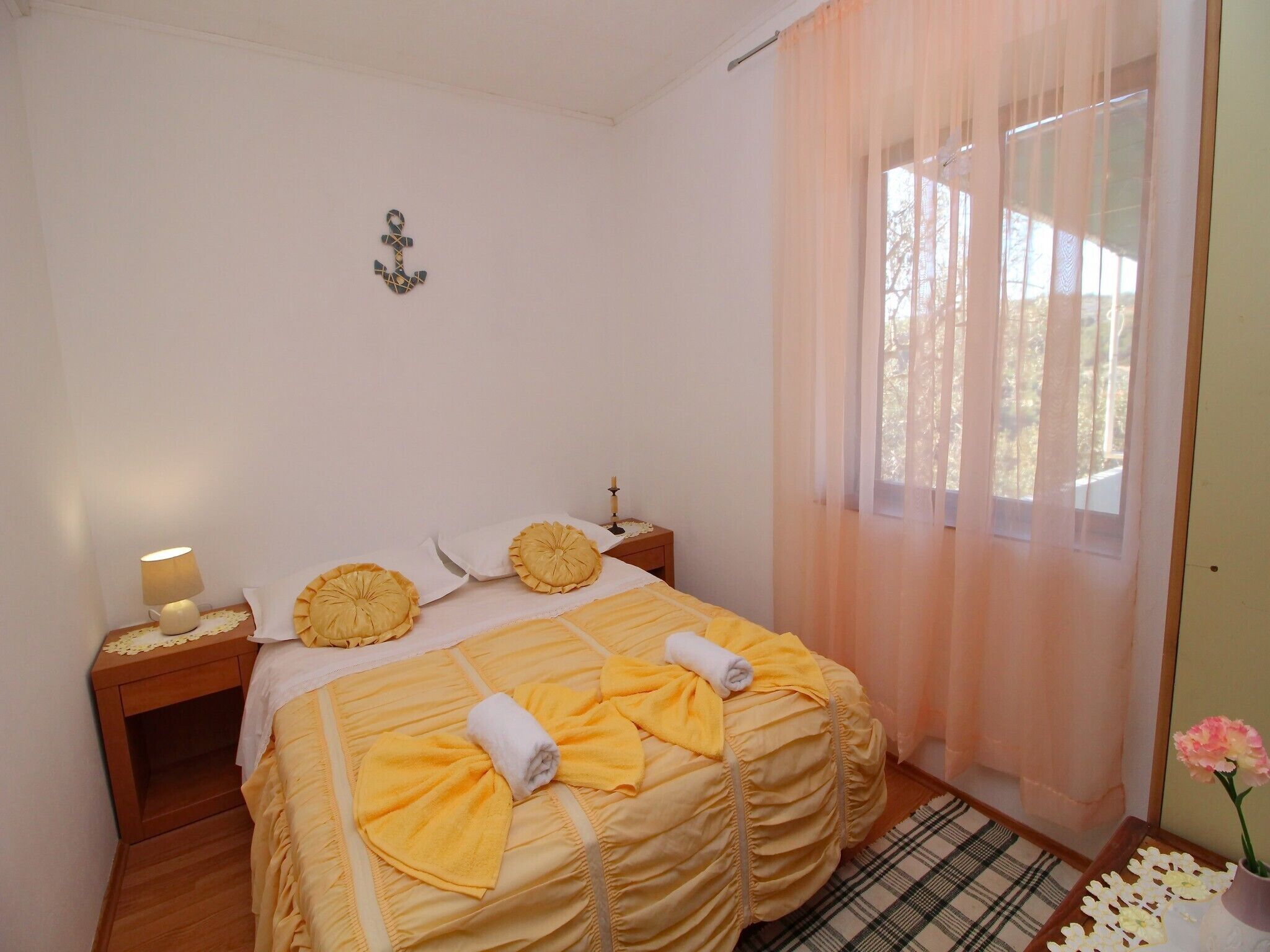 A neatly made bed with yellow linens and towels, a bedside lamp, a small nightstand, and a window with sheer curtains.