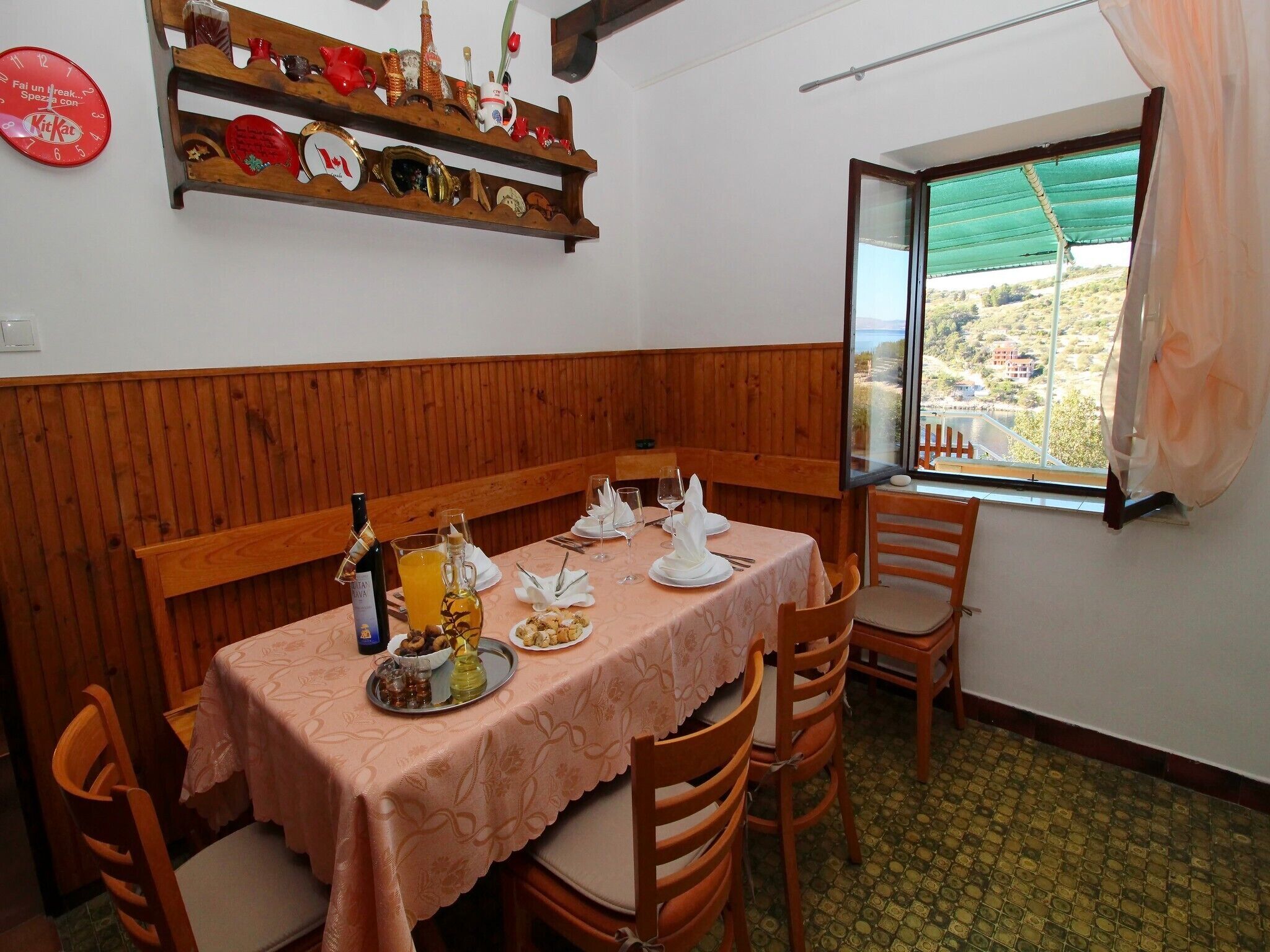 Un coin repas avec un banc en bois, une table dressée pour deux personnes avec des assiettes, des verres et une bouteille de vin. Une étagère située au-dessus contient divers objets décoratifs.
