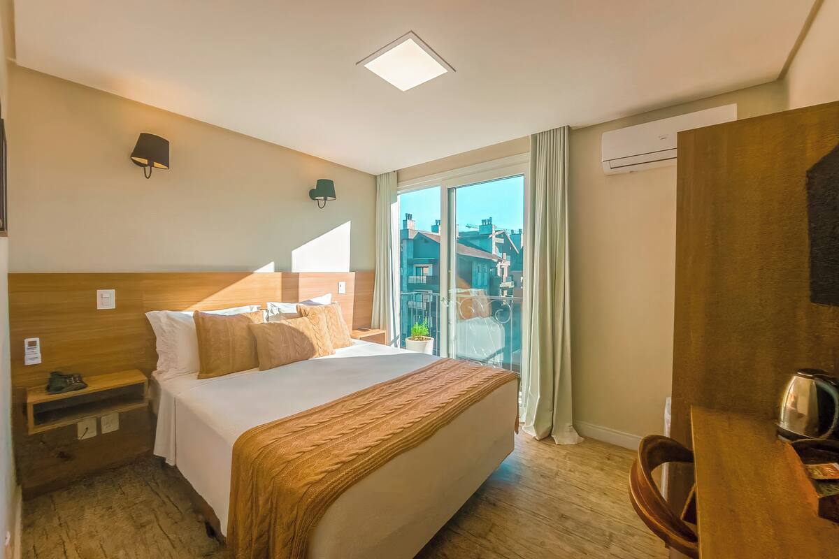 A hotel room with a bed, bedside table, lamp, TV, and a view of buildings through the window.
