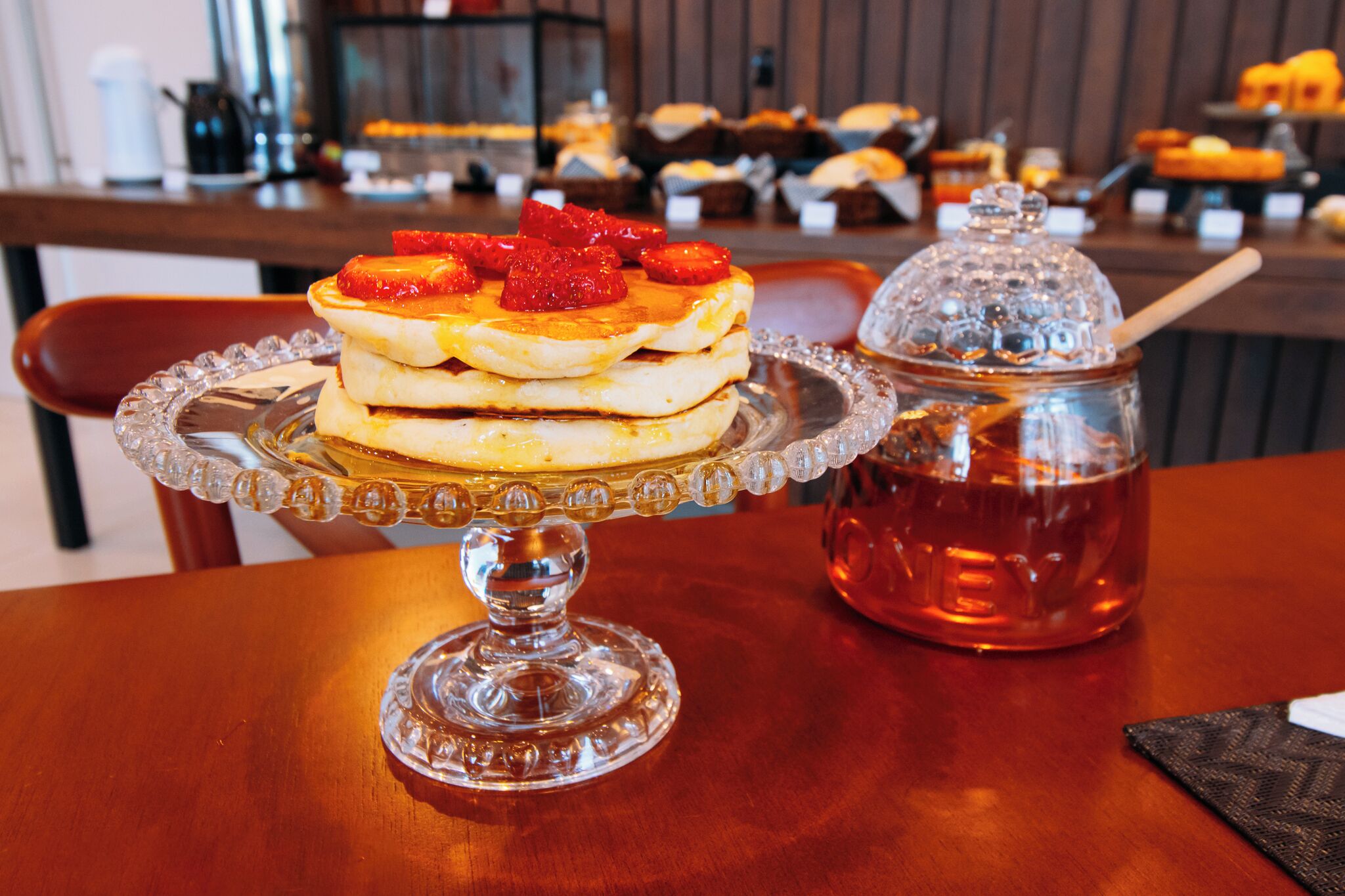 A breakfast setting with pancakes topped with strawberries and syrup, a glass pitcher, and a variety of pastries in the background.