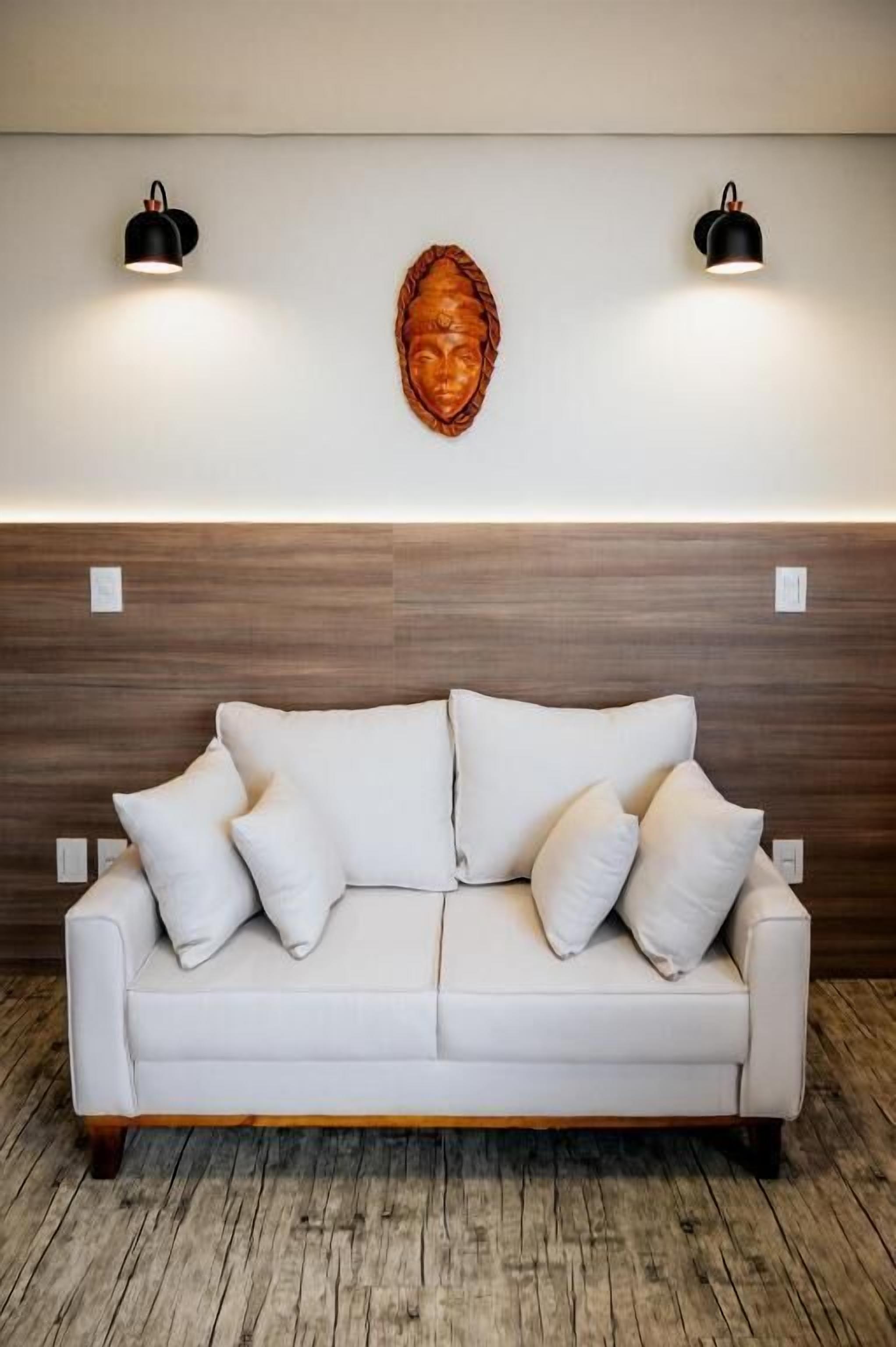 A white leather sofa with four cushions against a wooden headboard. Two wall-mounted lamps are on either side of a wooden wall art piece.