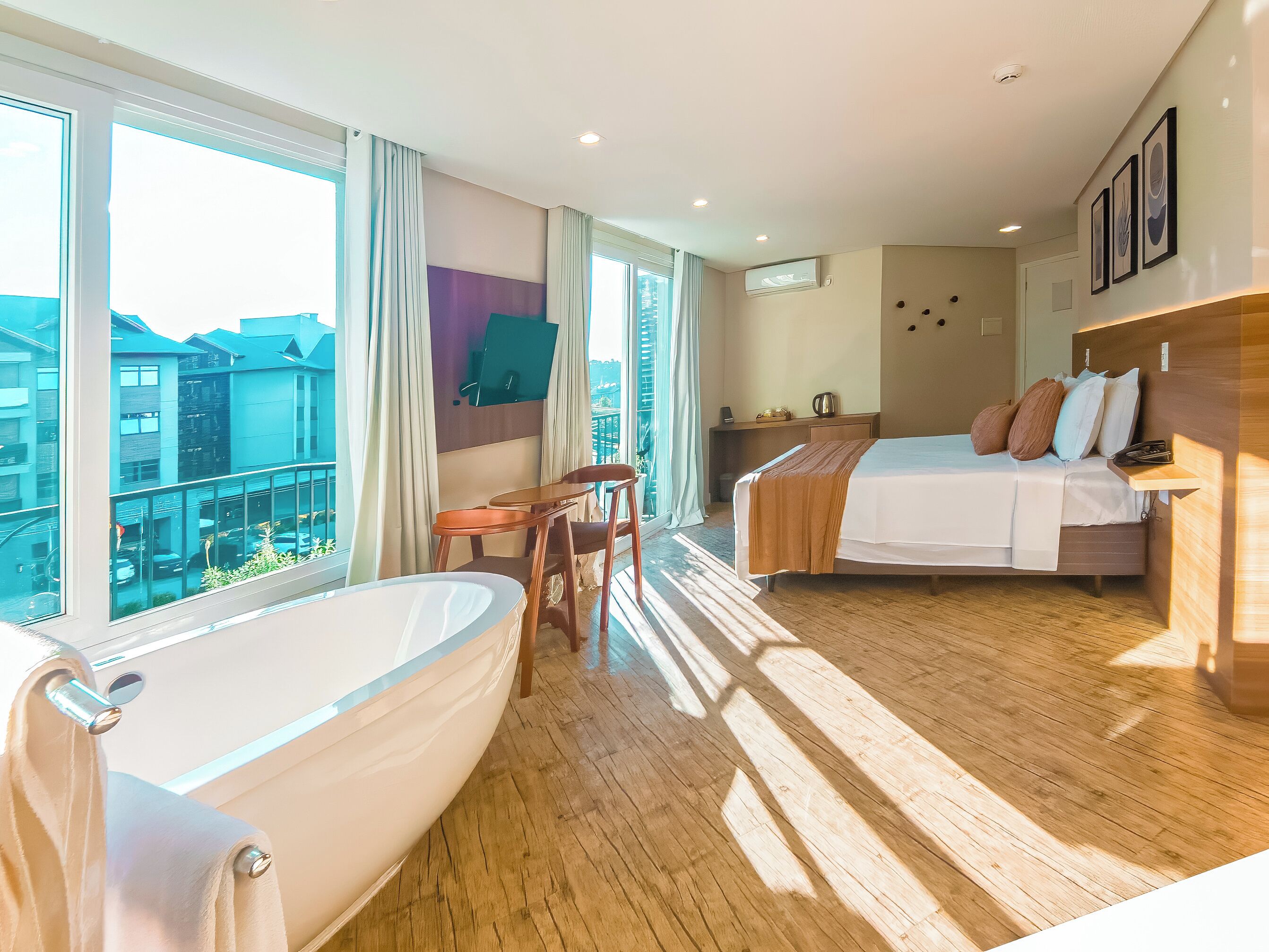 Een hotelkamer met een groot bad, een bed met veel kussens, een bureau met stoel, en uitzicht op gebouwen door grote ramen.