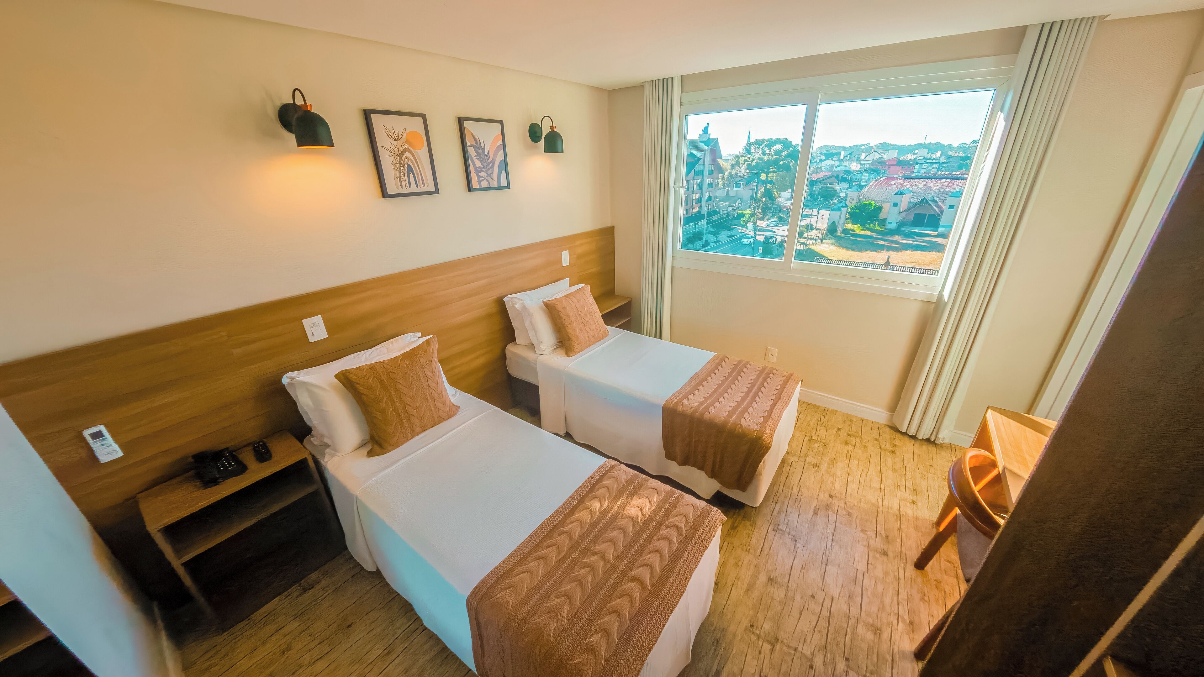 Quarto de hotel com duas camas, cabeceira de madeira, uma mesinha, uma cadeira e janela com vista para uma cidade.