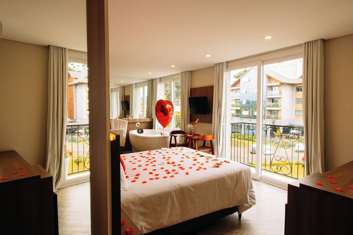 A hotel room with a bed, a bathtub, a TV, and a balcony with a view of buildings.