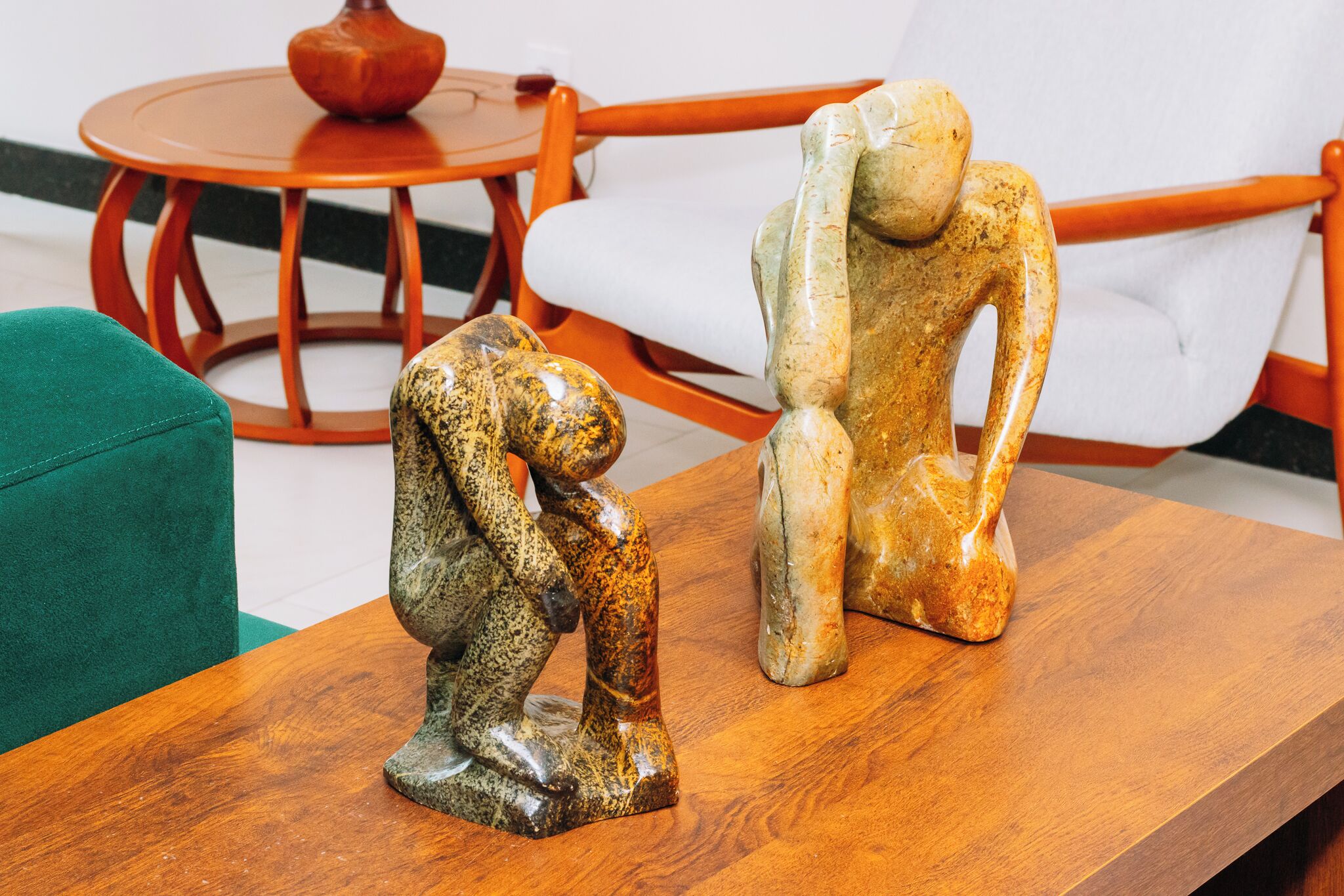 Two wooden sculptures on a table in a room with a green armchair and a round wooden table with a red vase.