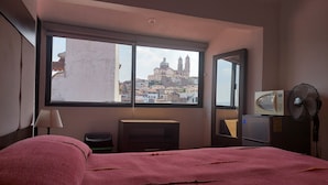 Habitación de hotel con un ventanal grande, una cama, un escritorio, una silla, un microondas y un ventilador.