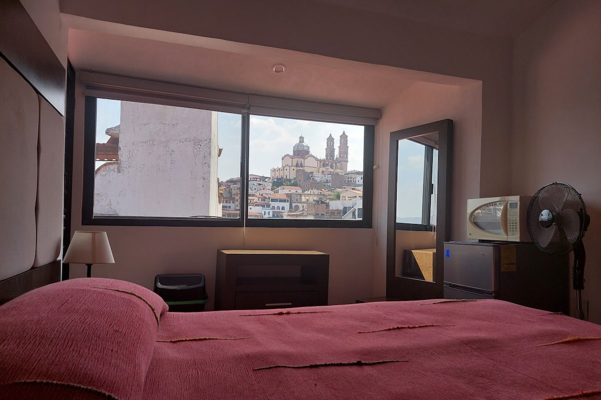 A hotel room with a large window, a bed, a desk, a chair, a microwave, and a fan.