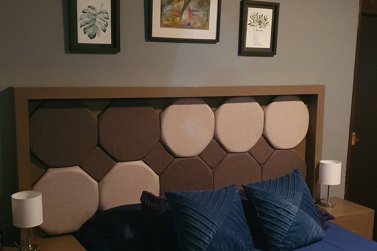 A bedroom with a large bed, two bedside tables, and a headboard with hexagonal panels. There are four framed artworks on the wall above the bed.