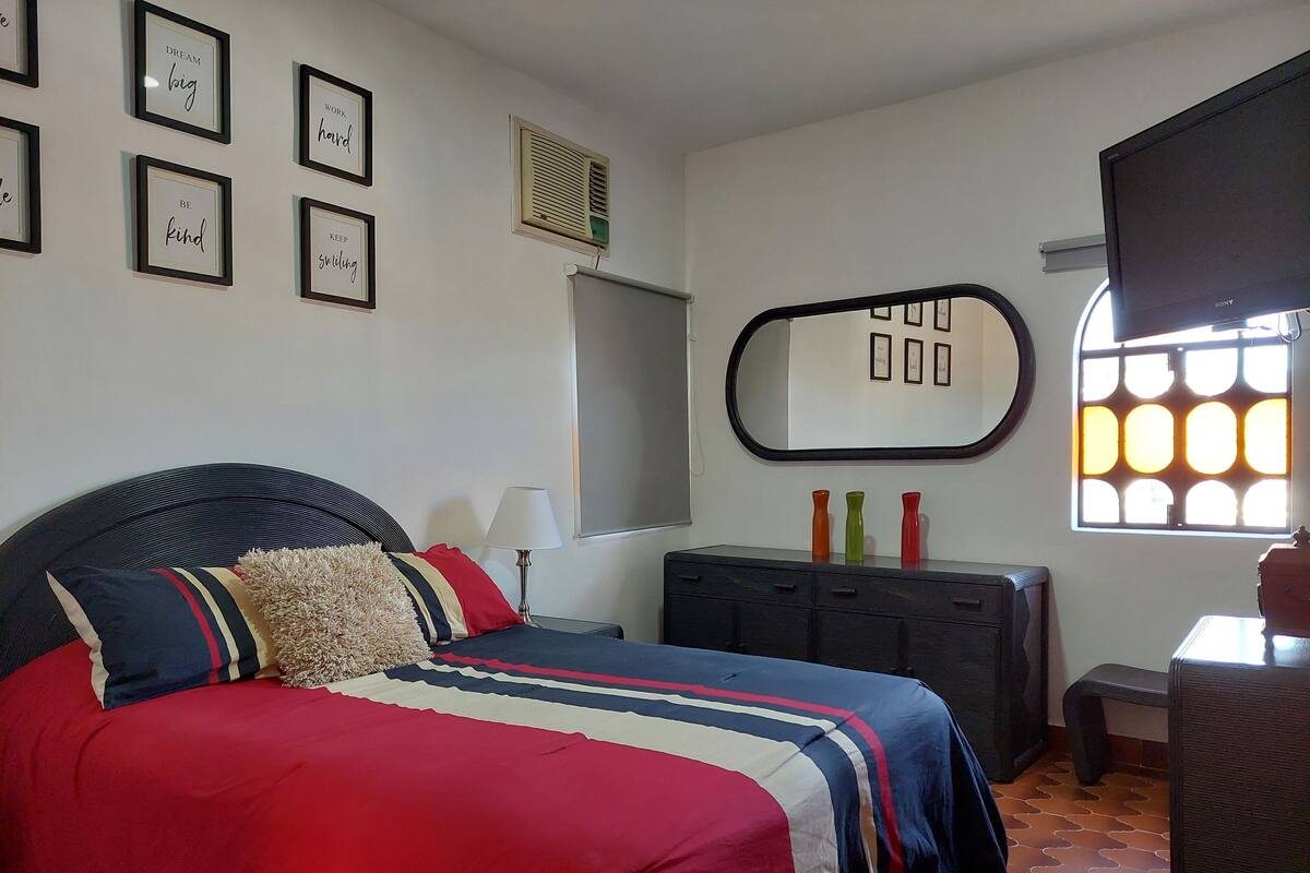 A bedroom with a bed, bedside table, lamp, dresser, framed motivational quotes, and a window with blinds.