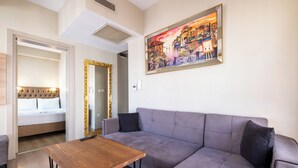 Una sala de estar con un sofá gris, una mesa de centro de madera y una cama con cabecera capitonada. Un cuadro grande enmarcados cuelga en la pared sobre el sofá.