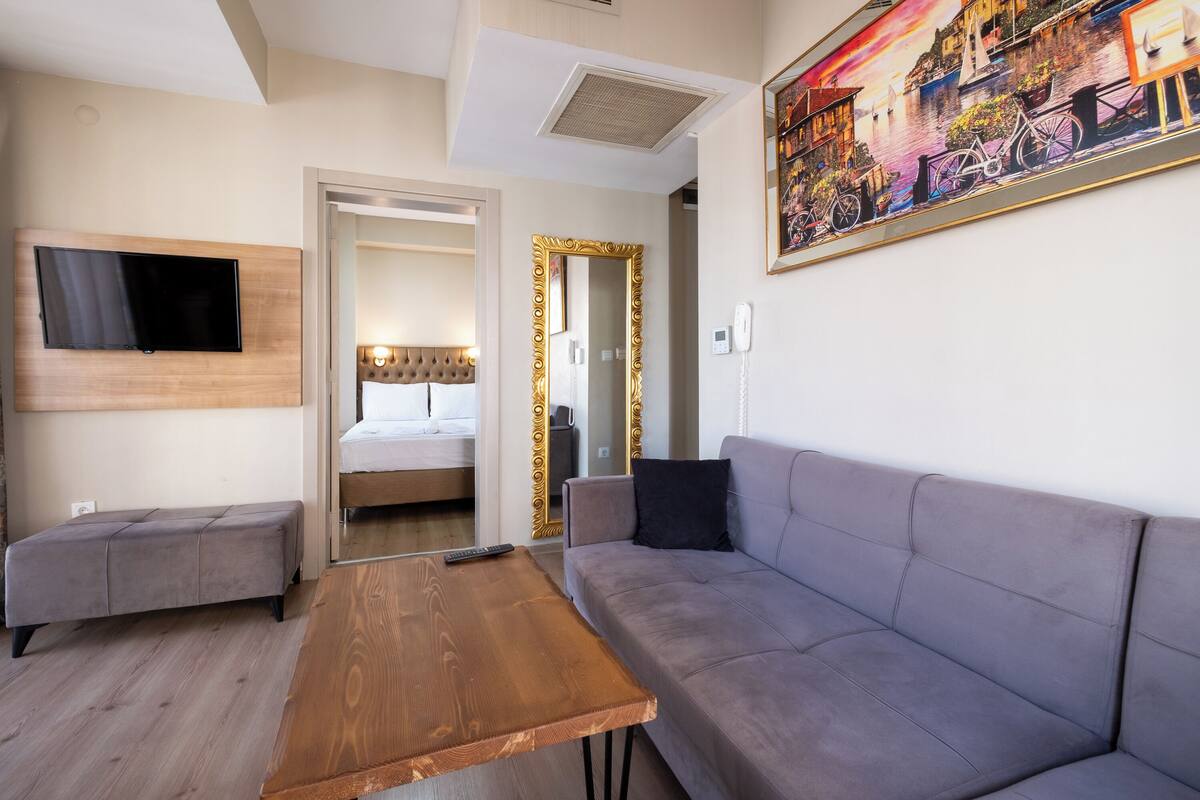 A modern hotel room with a grey sofa, a wooden coffee table, a flat-screen TV, and a painting of a coastal scene.