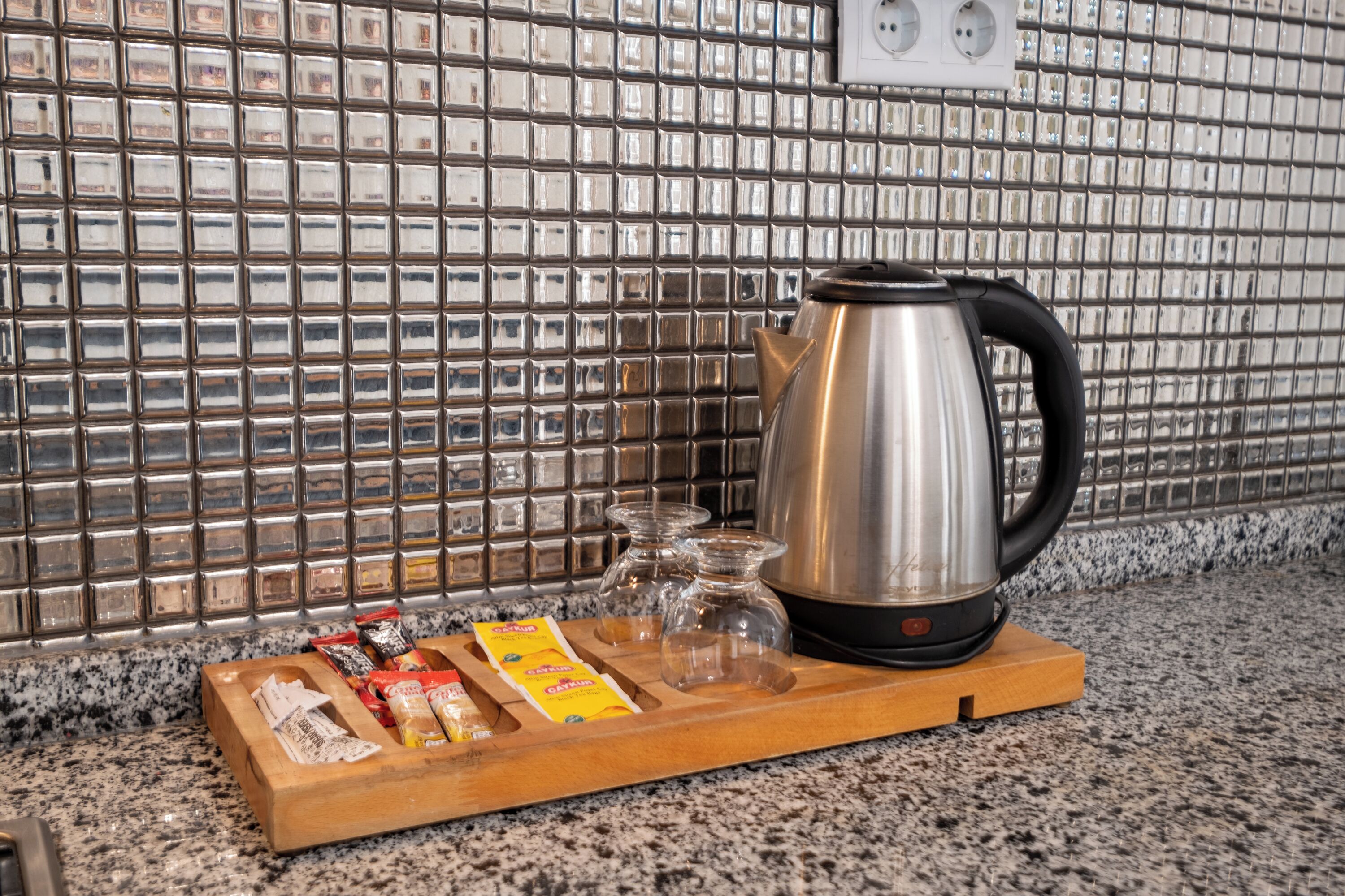 A kitchen counter with a stainless steel kettle, a wooden tray with packets, and clear glass cups.