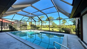 A swimming pool with a glass enclosure, surrounded by a metal frame and a glass roof, with a view of a green landscape and trees. - Osprey Preserve - spacious home overlooking Oscar Scherer Park (Nokomis)