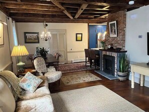Een woonkamer met een open haard, houten balken, een eethoek en een keuken die door een open deur zichtbaar is.