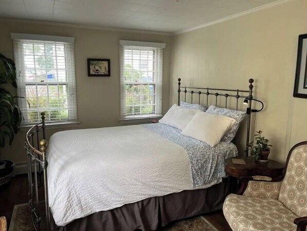 Une chambre à coucher avec un lit, deux fenêtres, une table de chevet, une chaise et un tableau au mur.