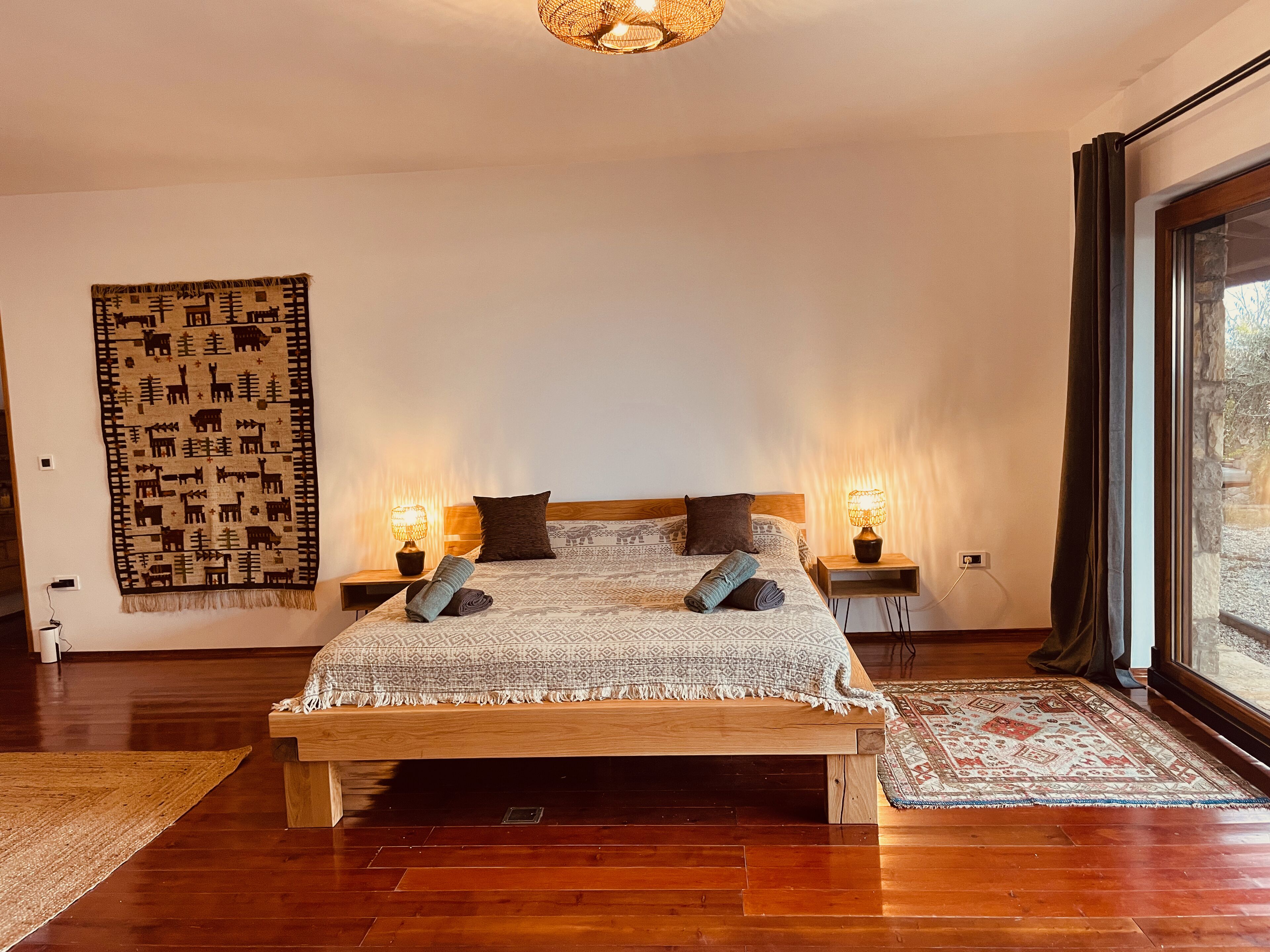 Une chambre à coucher avec un lit en bois, deux lampes de chevet, une grande tapisserie tissée et une vue par la fenêtre.
