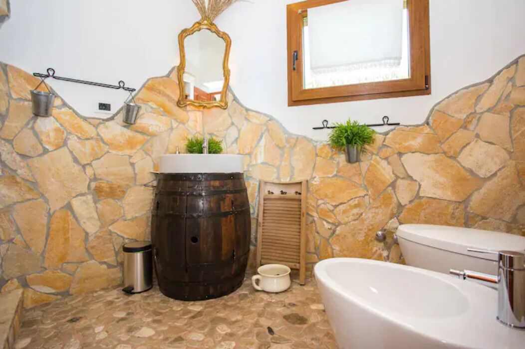 A bathroom with a stone wall, a wooden barrel, a white sink, a toilet, and a mirror.