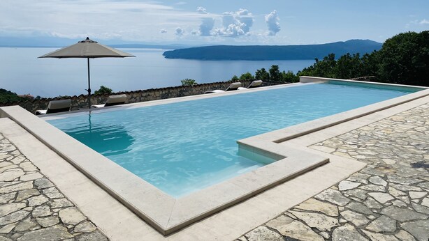 Una piscina infinita rectangular con borde de piedra, rodeada de sillones de descanso y un gran parasol, con vistas al mar tranquilo y montañas lejanas.