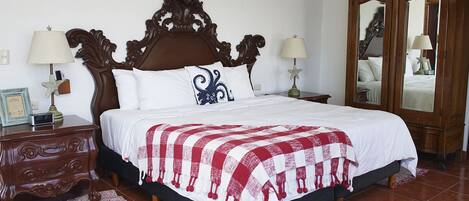 Una cama bien hecha con una colcha roja y blanca a cuadros, dos lámparas de noche, una mesita de madera con un diploma enmarcado y un mueble espejo grande.