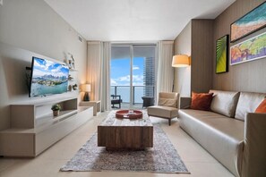 A modern living room with a large TV, a wooden coffee table, and a sofa with orange cushions. - ✨ 15th-Floor Modern 1BR @ Lyfe Resort | Ocean View + City Skyline | Luxury Amenities (Hollywood)