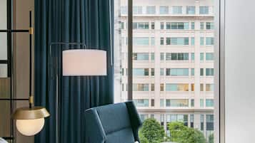 Une chambre d’hôtel avec un lit, un fauteuil bleu, une lampe sur pied, une vue sur un immeuble et un rideau.
