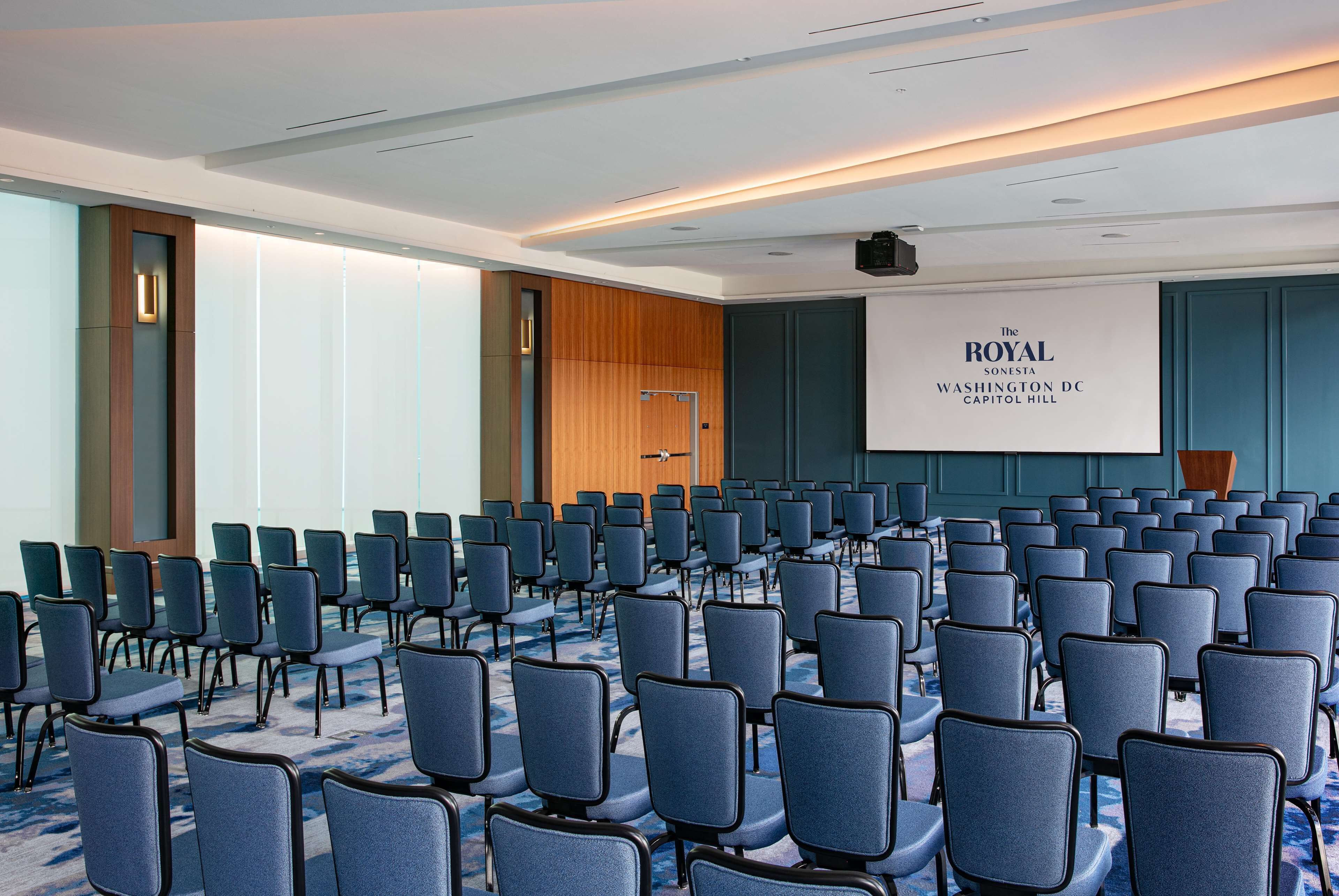 A conference room with rows of chairs, a podium, and a screen displaying "The Royal Sonesta Washington DC Capitol Hill".