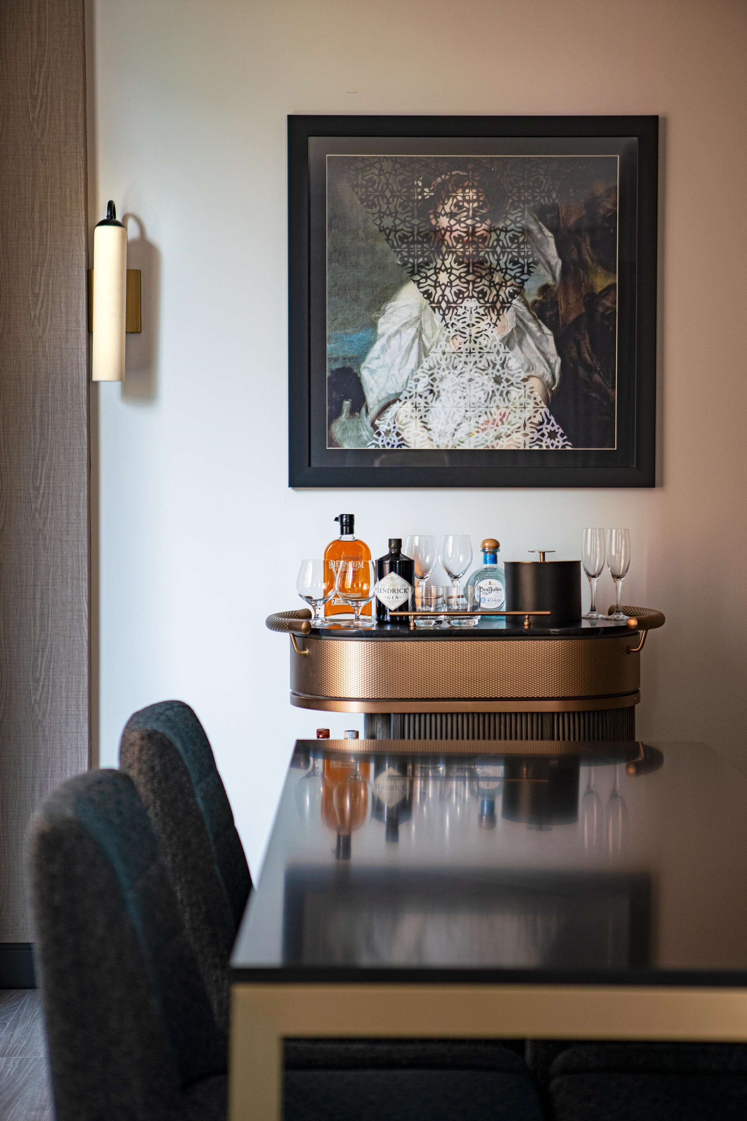 A dining table with a tray of bottles and glasses, a framed artwork above, and a lamp on the wall.