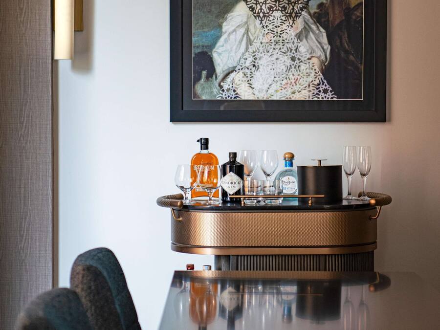A dining table with a tray of bottles and glasses, a framed artwork above, and a lamp on the wall.