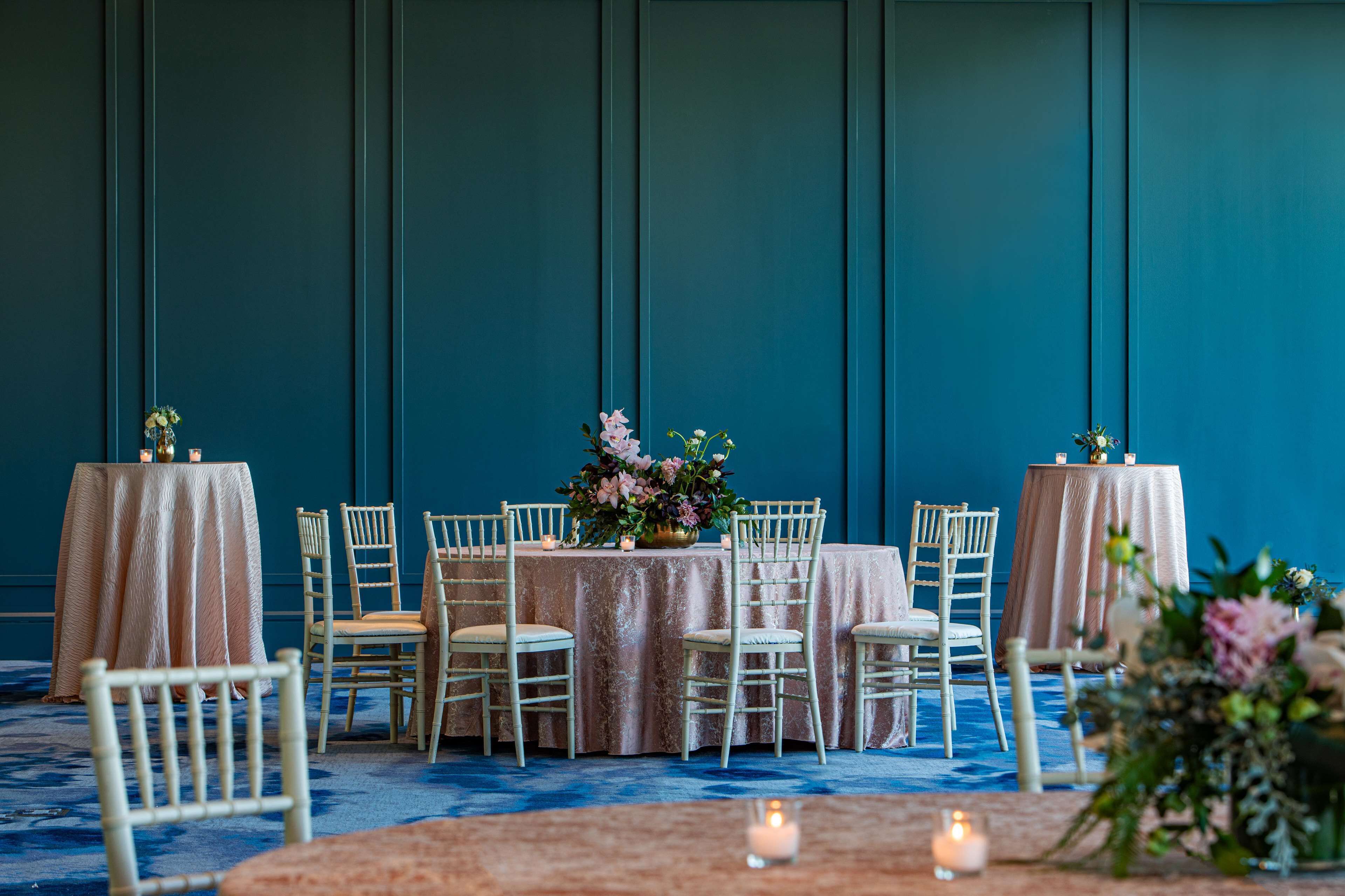 A room with a blue wall, round tables with white chairs, and floral centerpieces.