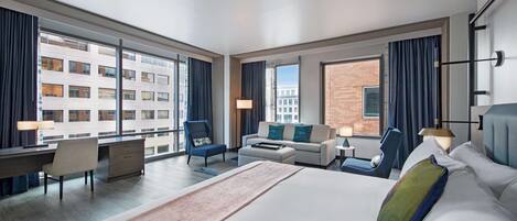 A modern hotel room with a large bed, a desk, a sofa, and a chair. There are large windows with curtains, and the room has a view of a building outside.