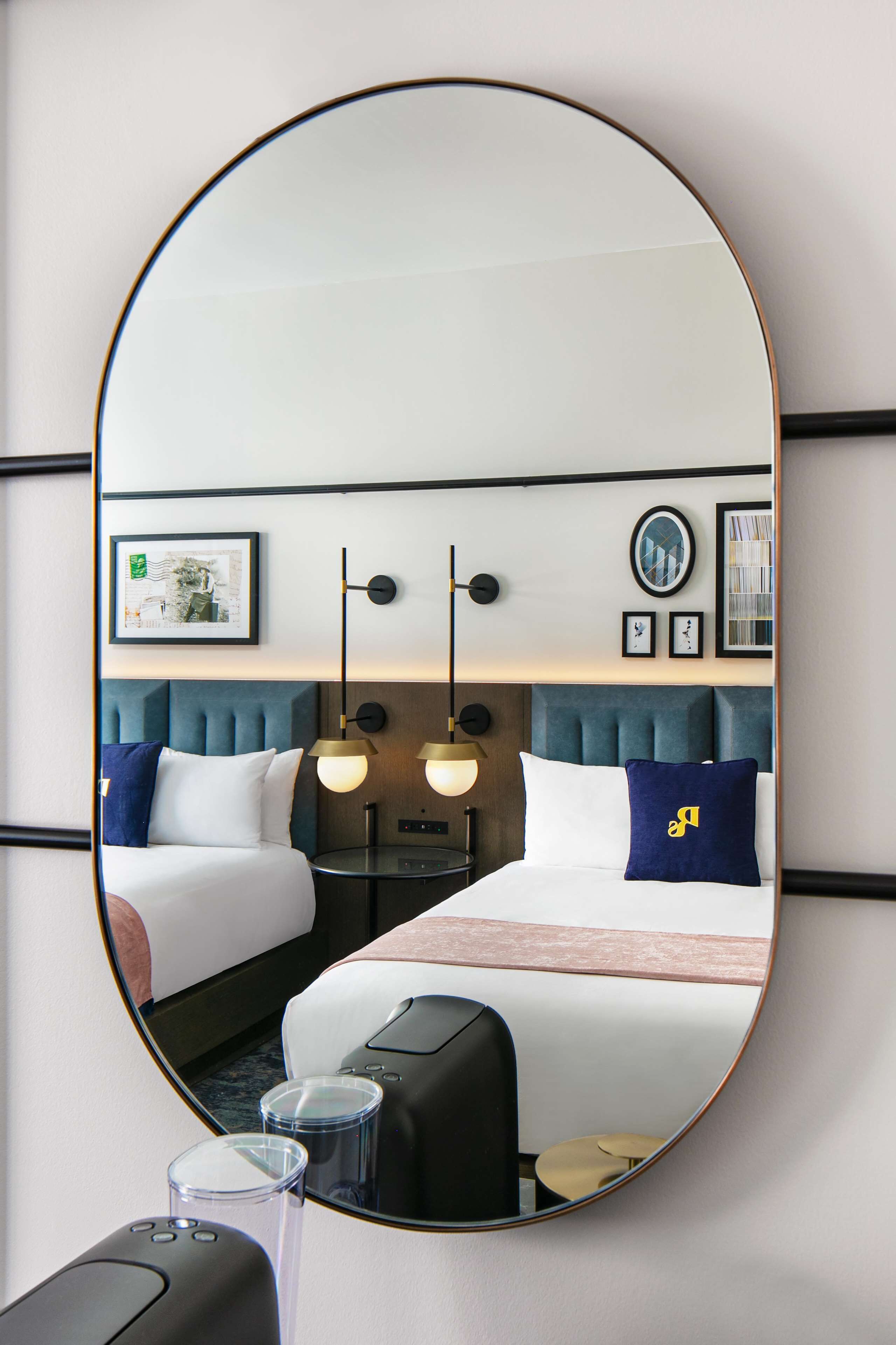 A hotel room with a large oval mirror reflecting the bed, bedside tables, and wall decor.