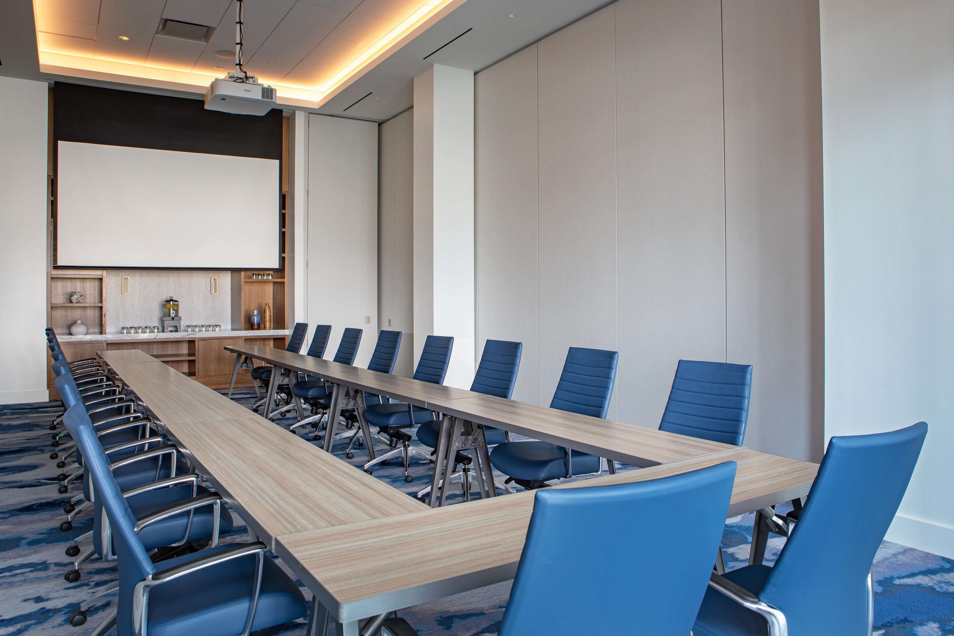 A conference room with a long table, blue chairs, a projector, and a white screen.