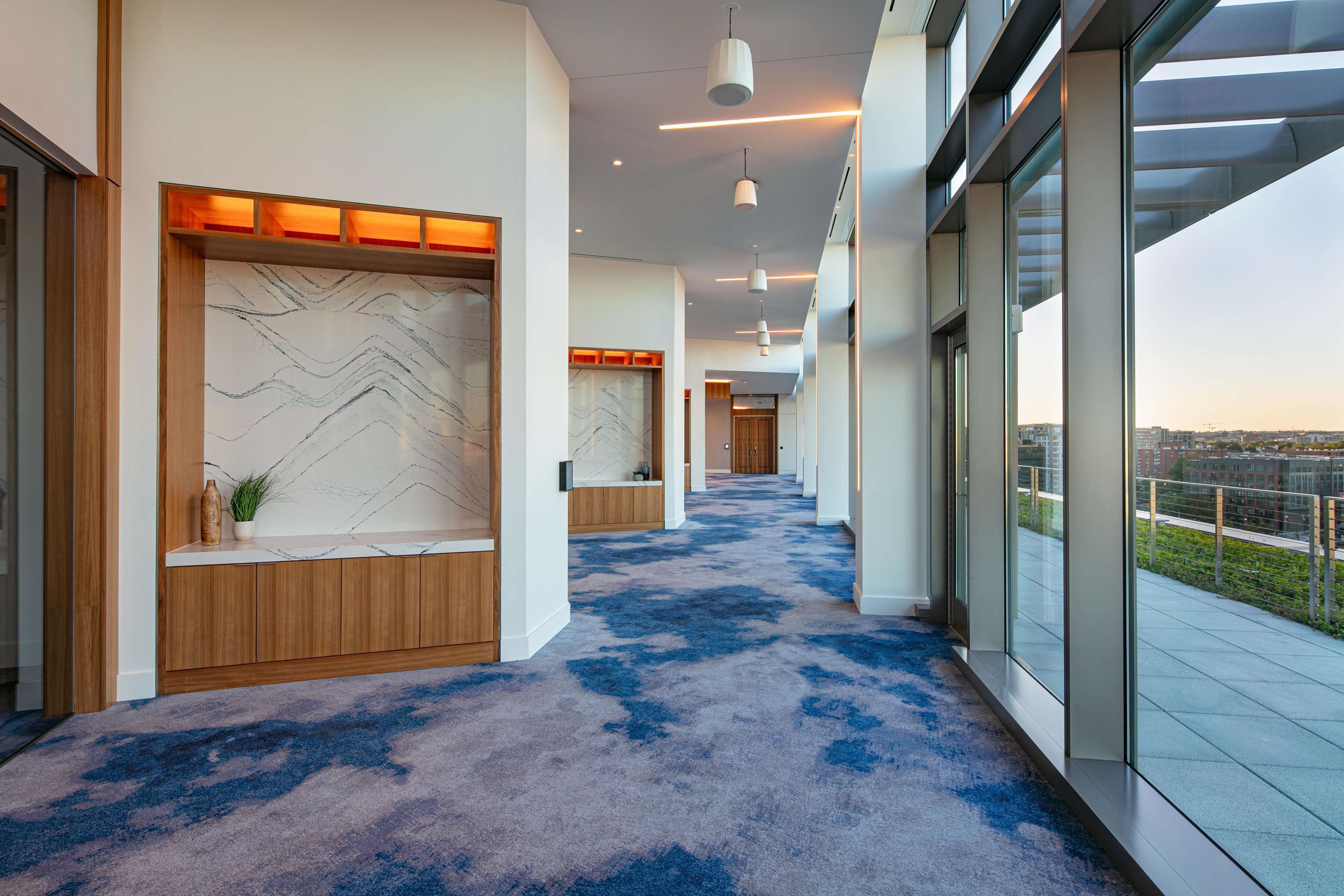A modern hotel corridor with a blue carpet, wooden reception desk, and large windows offering a city view.