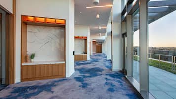 A modern hotel corridor with a blue carpet, wooden reception desk, and large windows offering a city view.