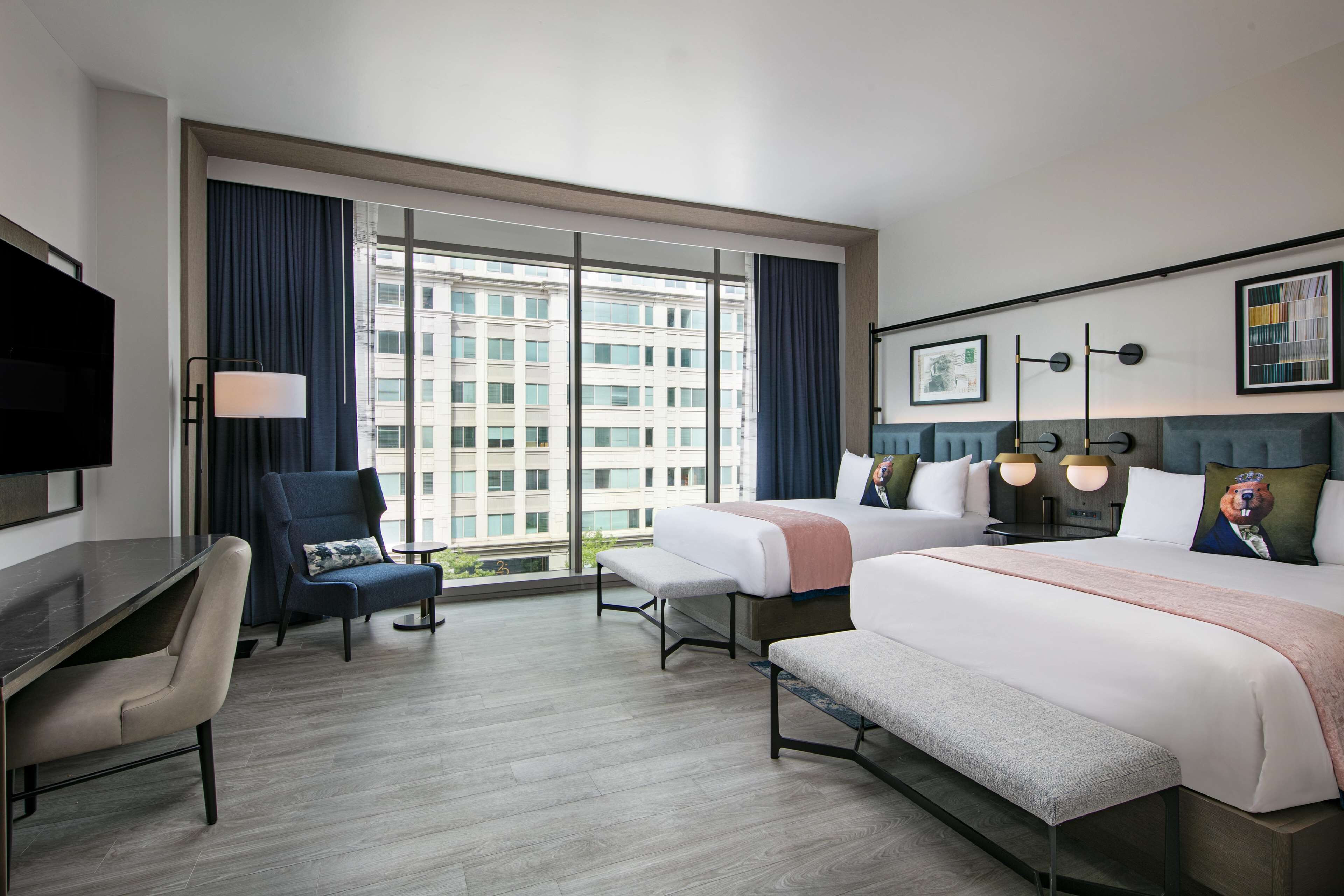 A hotel room with a large window, a desk, two beds, and a bench.