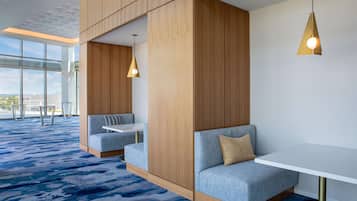 A modern hotel room with a large window, a blue carpet, a white table, and a blue sofa with a gold lamp.