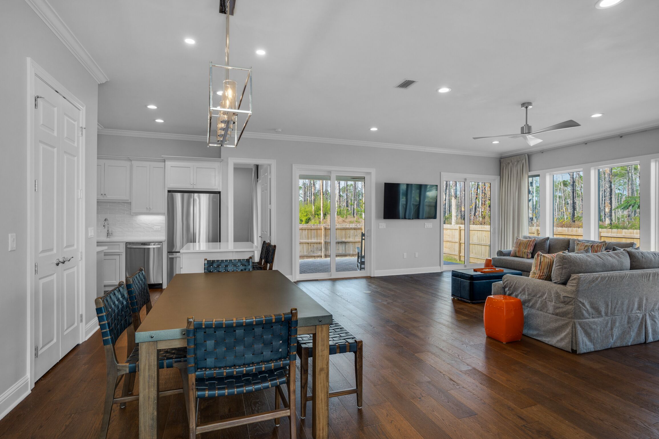 A spacious kitchen with white cabinets, a large wooden dining table, and blue chairs. There is a sliding glass door leading to a patio area with a gray sofa and orange ottoman.