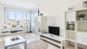 Uma sala de estar clara e moderna, com uma televisão grande, móveis brancos e um espaço de refeições com mesa e cadeiras.