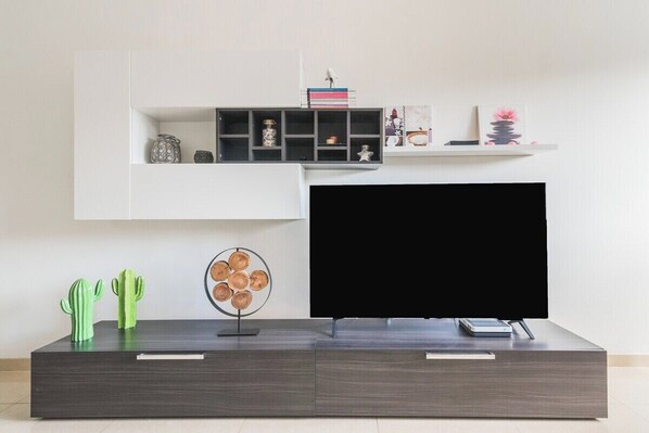 Una sala de estar moderna con un televisor de pantalla plana, mueble de entretenimiento de madera oscura y un estante blanco sobre el televisor con objetos decorativos.