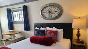 A neatly made bed with a red blanket, a headboard with decorative pillows, a round wall decoration, a small table with a lamp, and a window with curtains. - Cabin 21 - Bicycle Retreat (Helena)