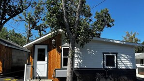 Ett enplanshus på hjul med vit fasad och en träpanel på vänster sida. Det finns en vit dörr med ett fönster, en liten veranda med en bänk och ett träd framför huset.
